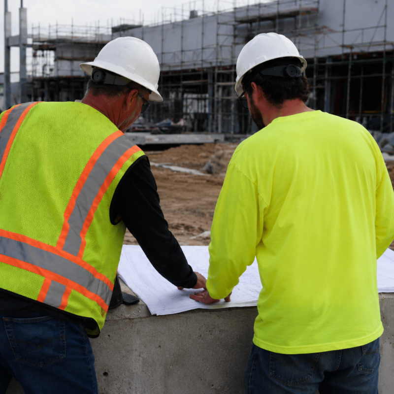 Construction project manager reviewing job costing data on a tablet at an active job site with crews working in the background.