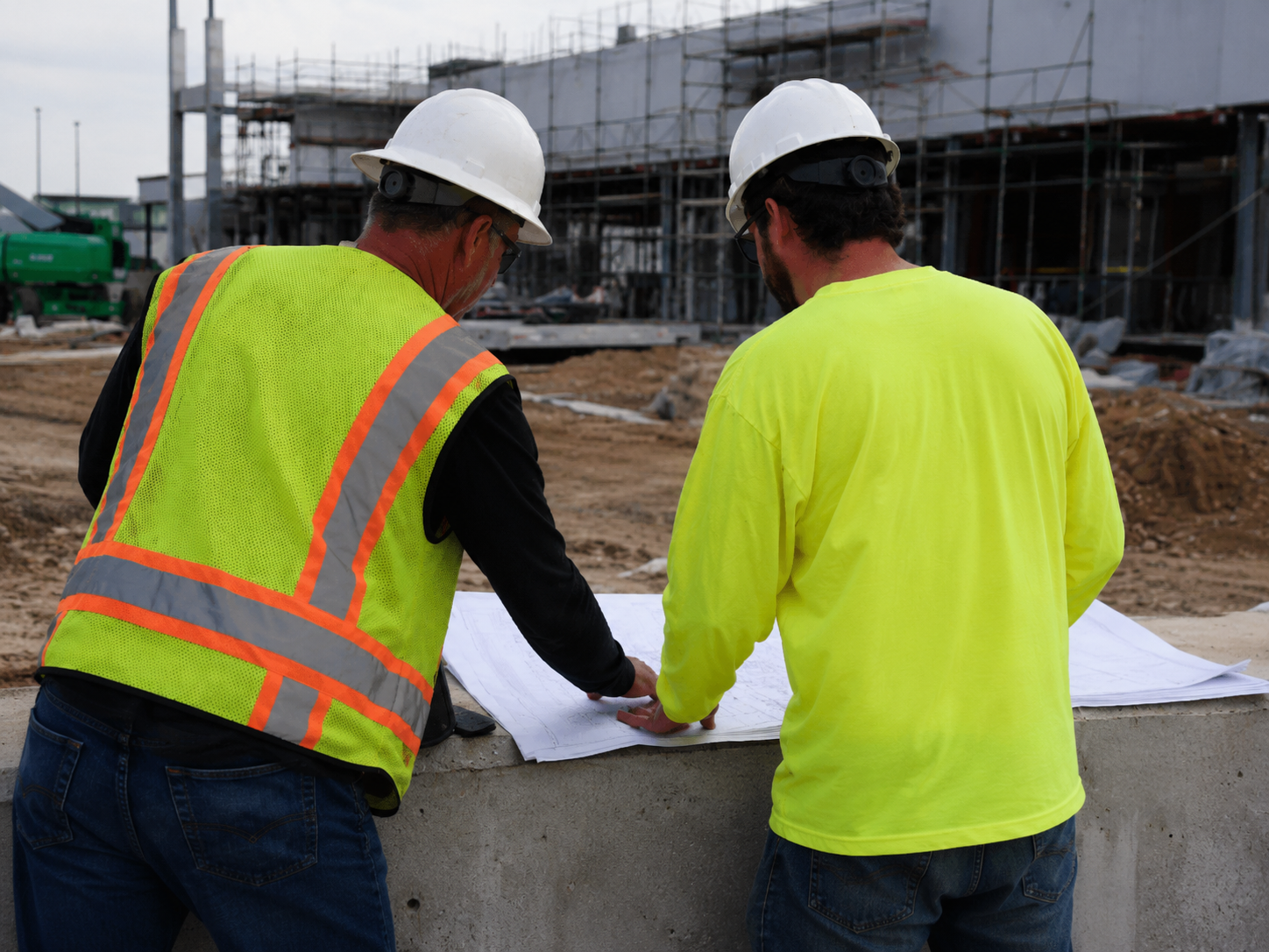 Construction project manager reviewing job costing data on a tablet at an active job site with crews working in the background.