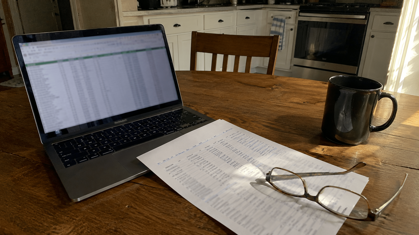 A home kitchen table at dawn with a laptop, a printed spreadsheet with illegible numbers, coffee and reading glasses (no text, no faces, no logos)