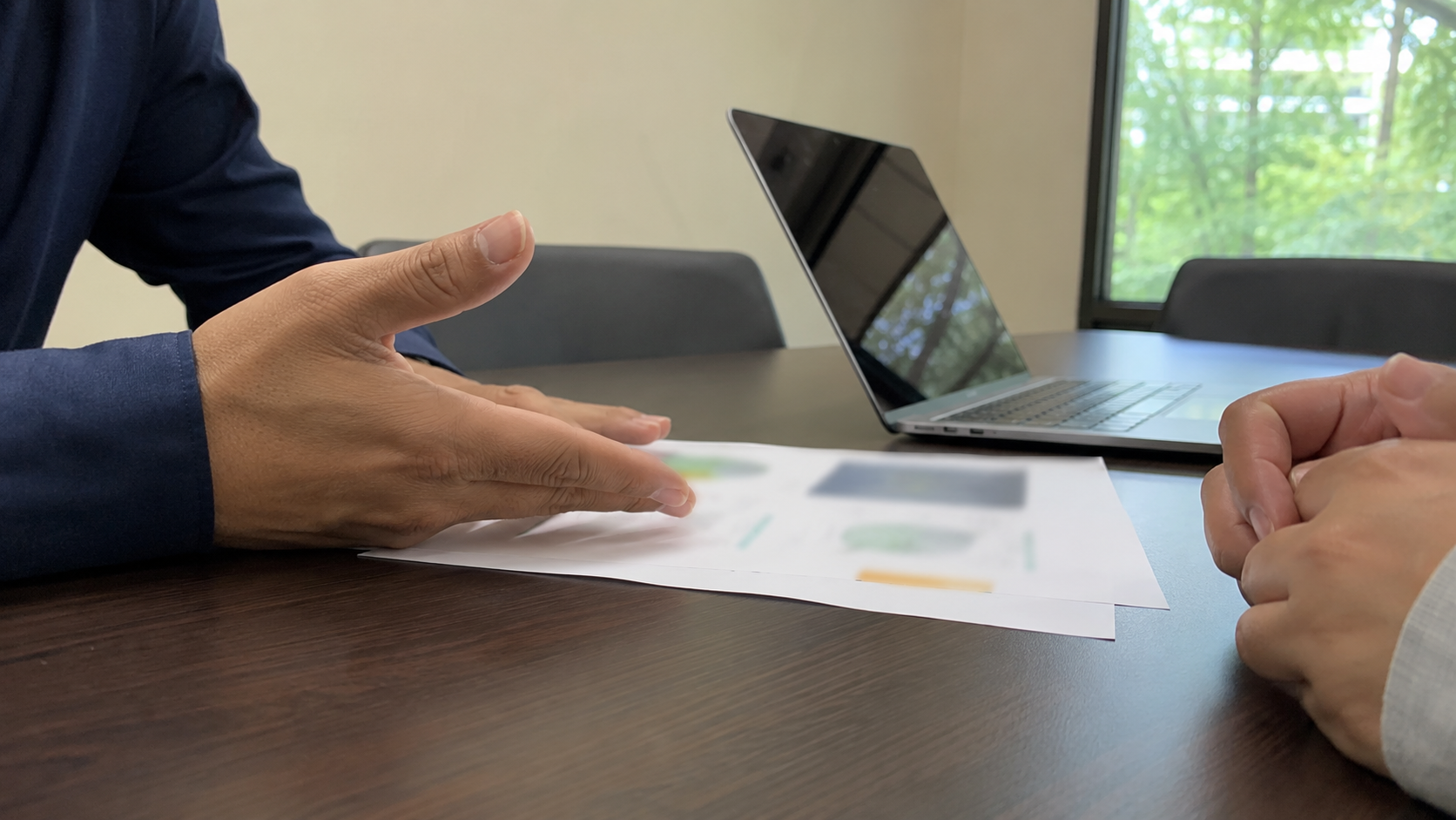 Two people's hands across a small meeting table, one gesturing over a printed report with illegible figures, a laptop nearby (no faces, no logos)