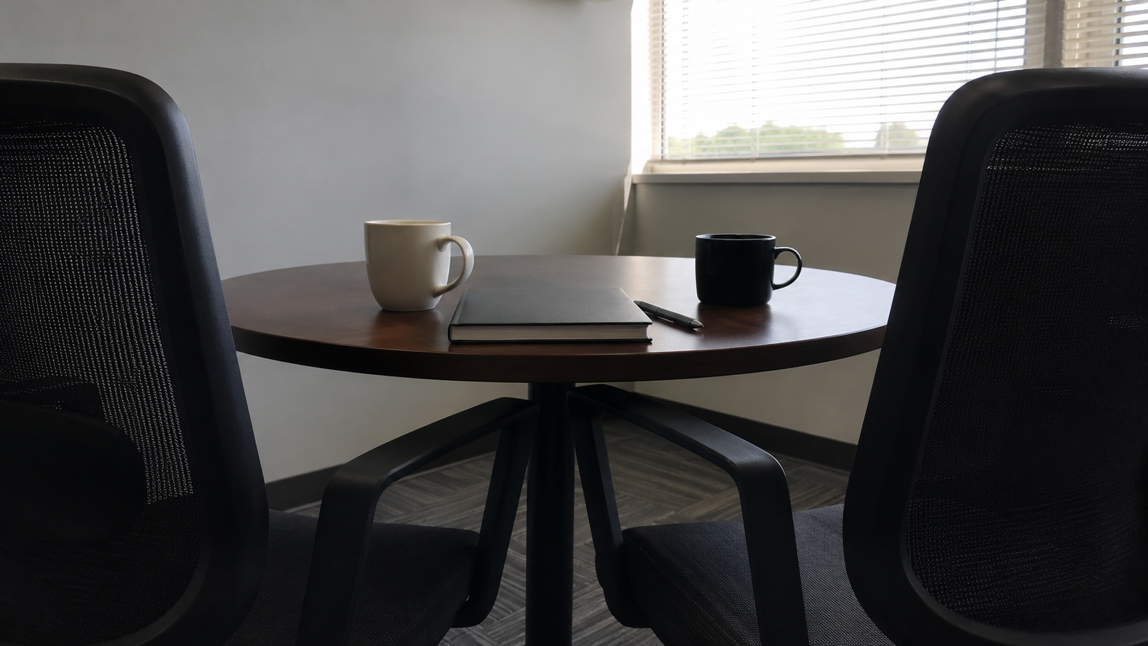 Two empty chairs at a small round office table with a blank closed notebook, two coffee cups and a pen, in half-open-blind morning light, no faces or logos.