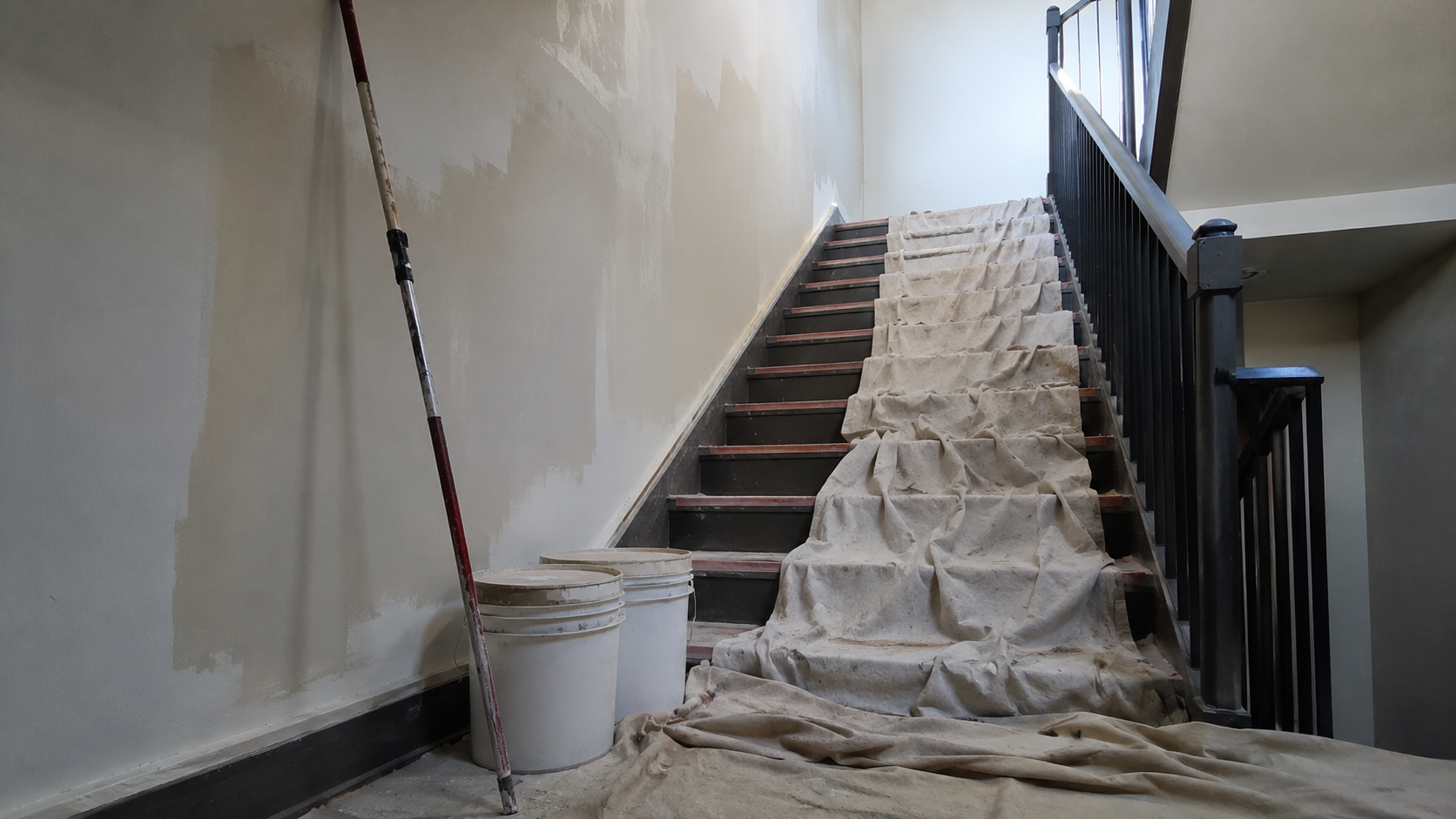 A partially painted commercial stairwell with drop cloths, a roller pole and two unlabeled paint buckets, in flat midday light, no faces or logos.