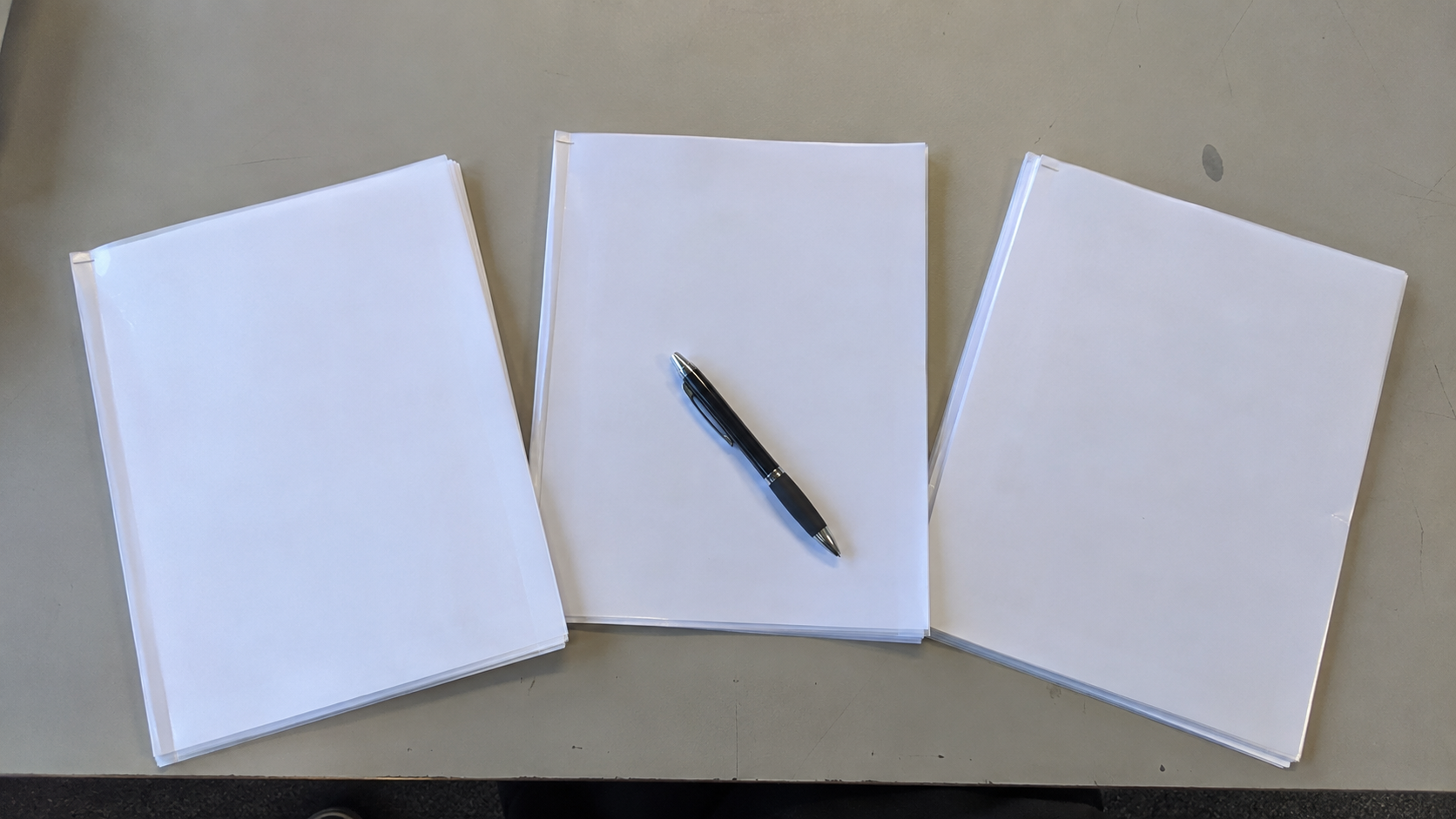 Three printed proposal packets fanned side by side on a plain desk with pages angled so no text is legible and a pen resting across the middle one, no people or logos.