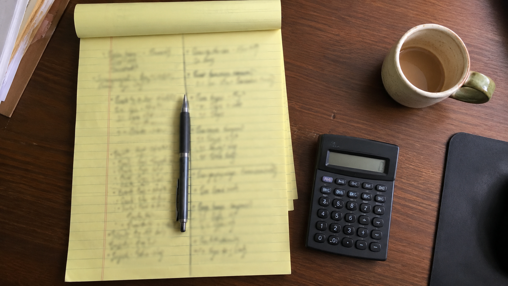 A legal pad with two handwritten columns of illegible figures, a calculator and coffee, shot top-down (no text legible, no logos)