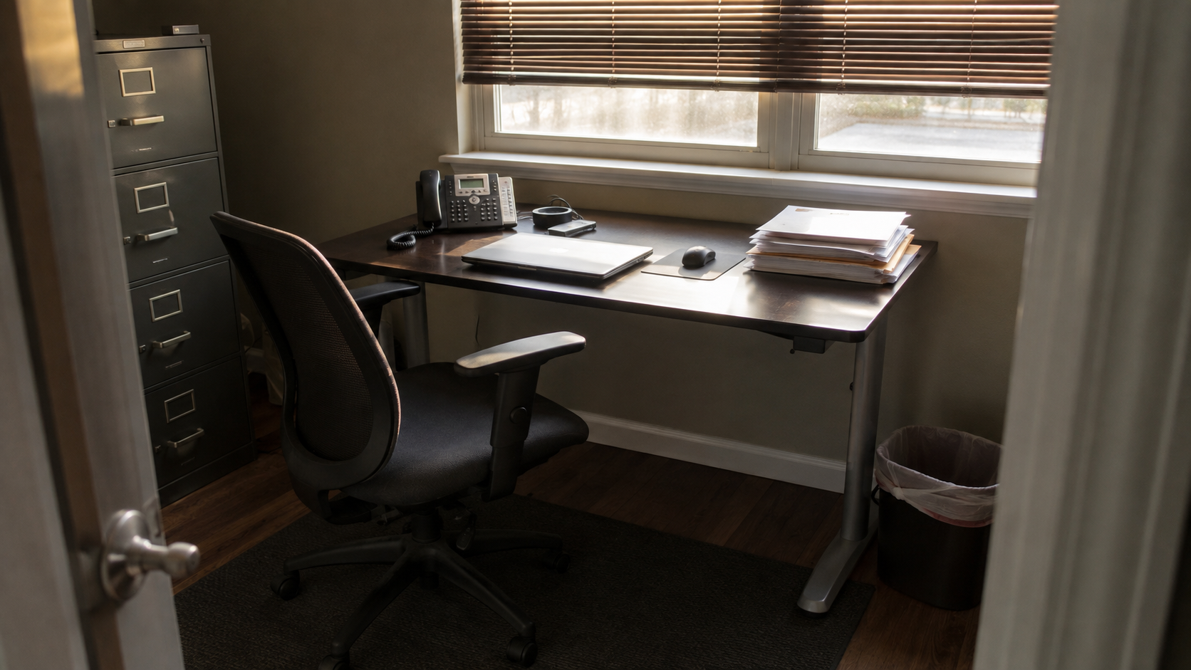An empty office chair pushed back from a small contractor back-office desk with a closed laptop and unopened mail