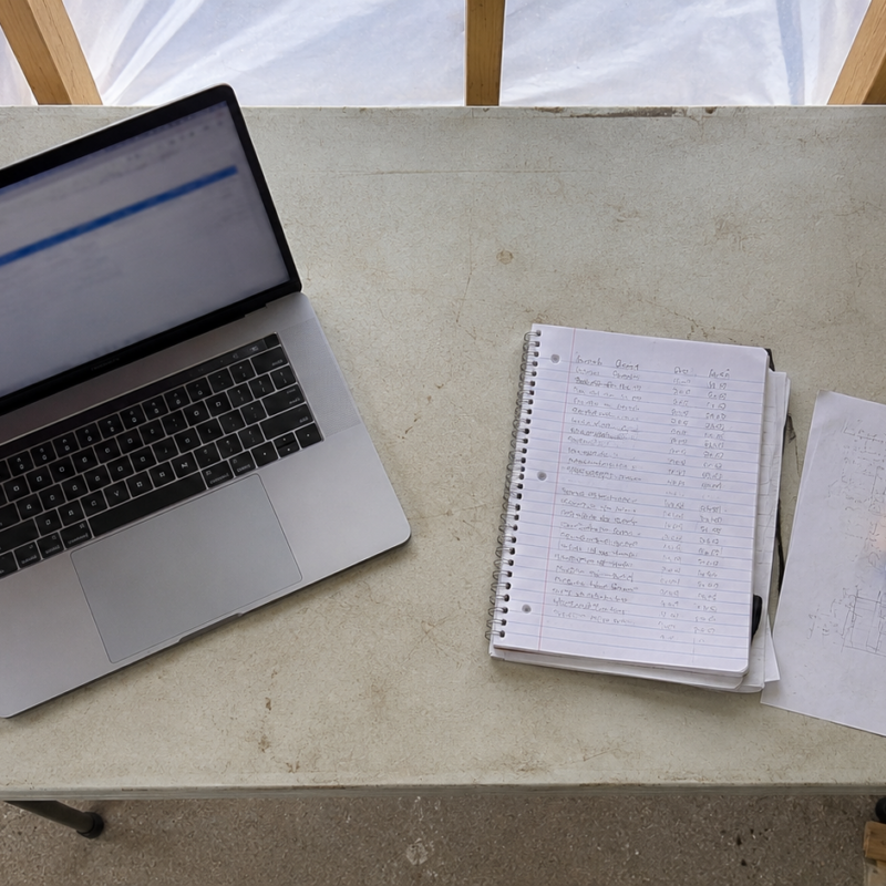 A laptop and a handwritten notebook of figures on a folding table inside a building under construction