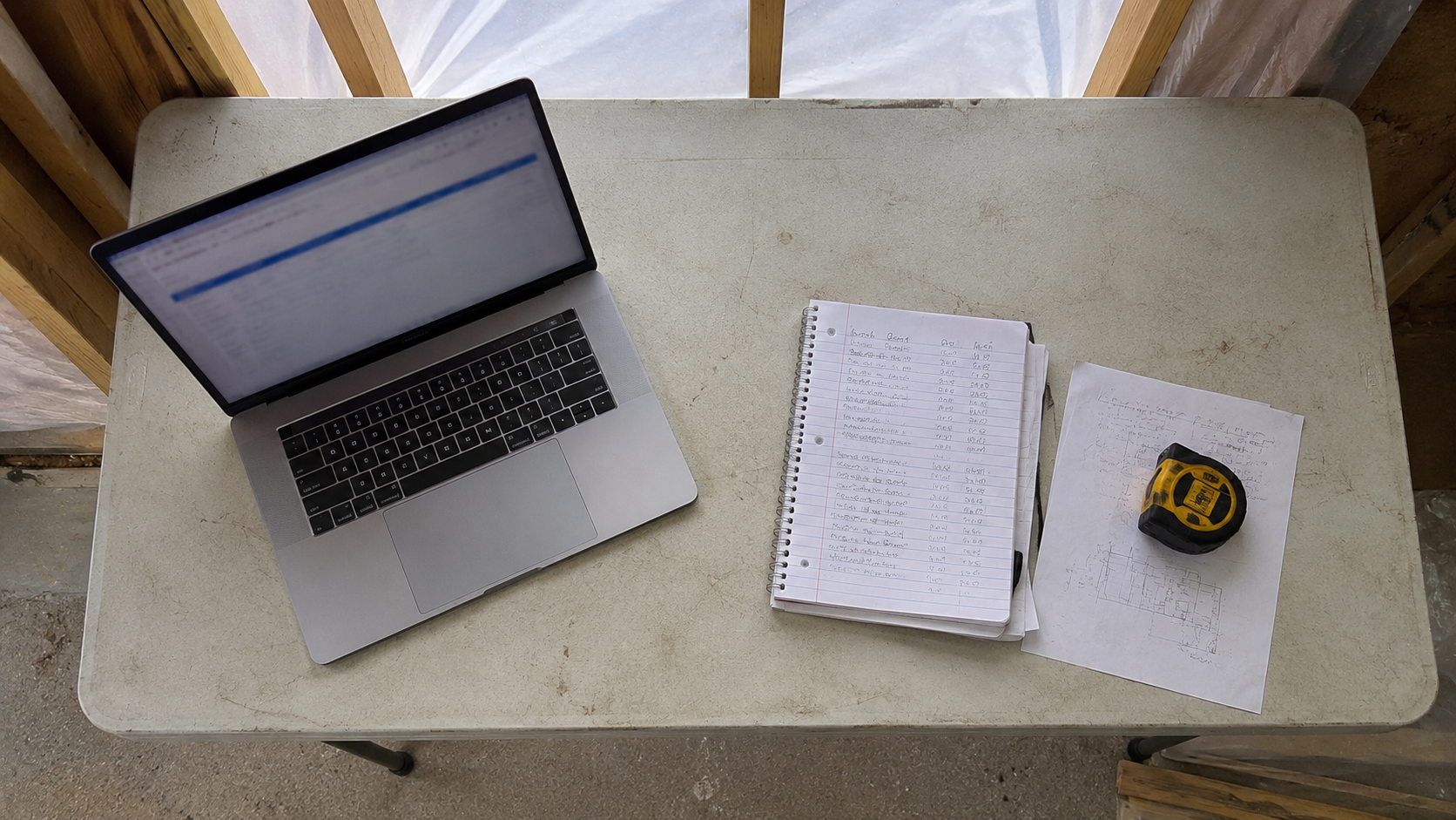 A laptop and a handwritten notebook of figures on a folding table inside a building under construction