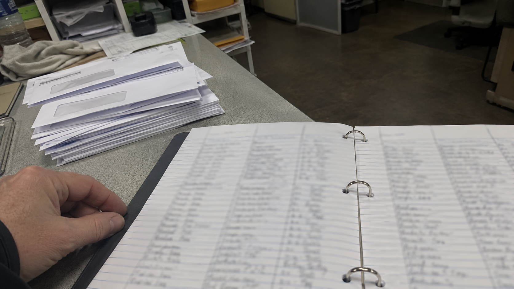 A three-ring binder of handwritten entries with illegible writing on a trades back-office bench beside a stack of unopened envelopes, under fluorescent light, no faces or logos.