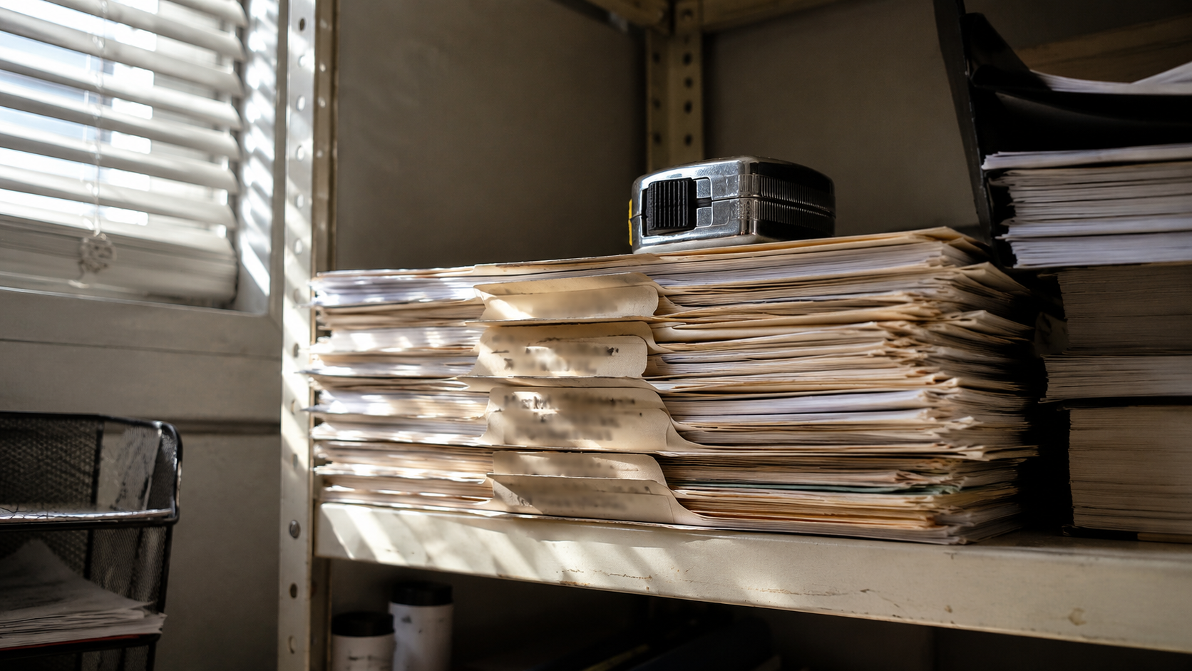 A stack of worn manila job folders with illegible handwritten tabs on a contractor office shelf under slatted blind shadows, with a tape measure on top, no people or logos.