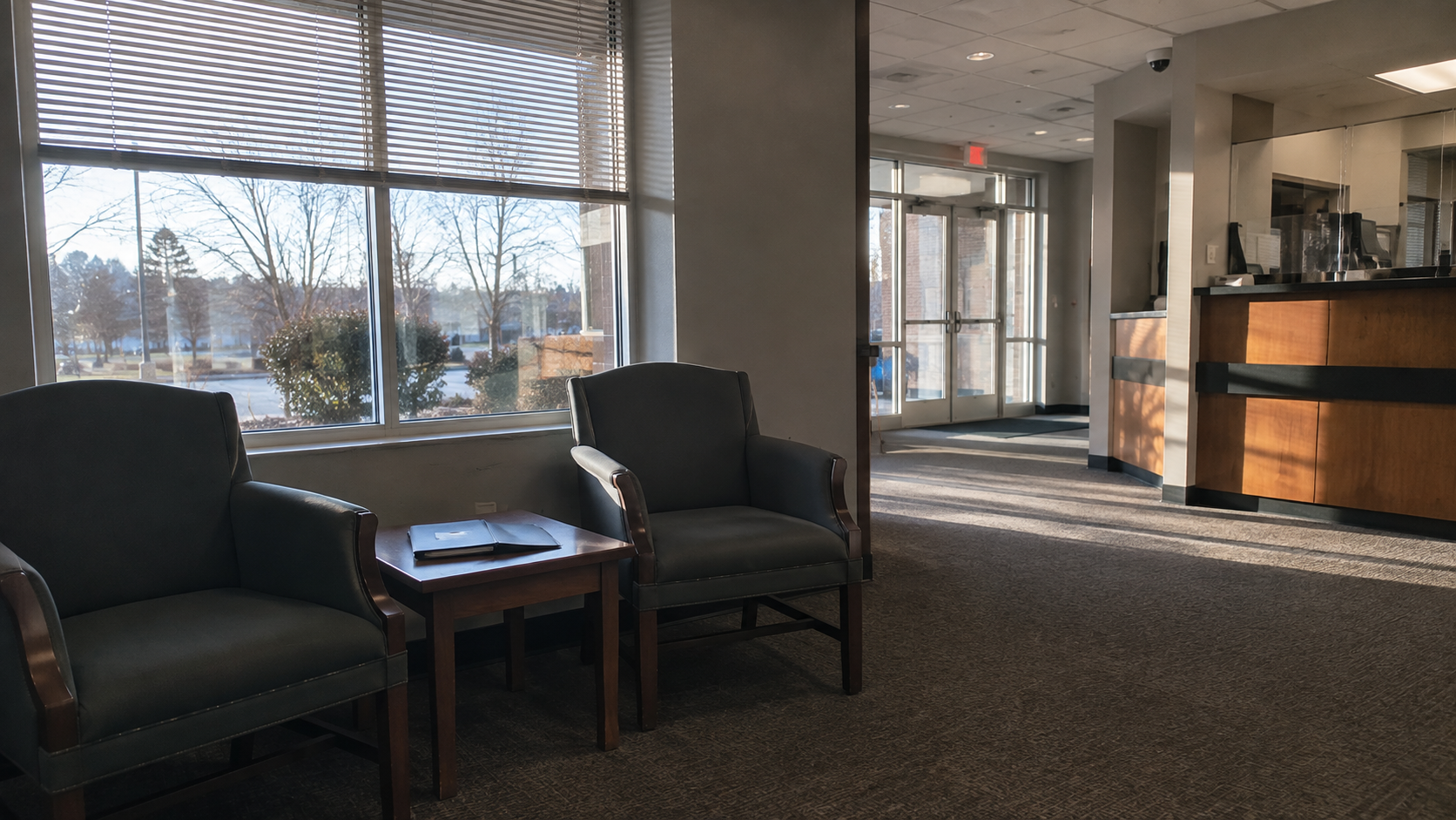 Two chairs and a small table with a closed folder by a window in an empty bank lobby in morning light, no people or logos