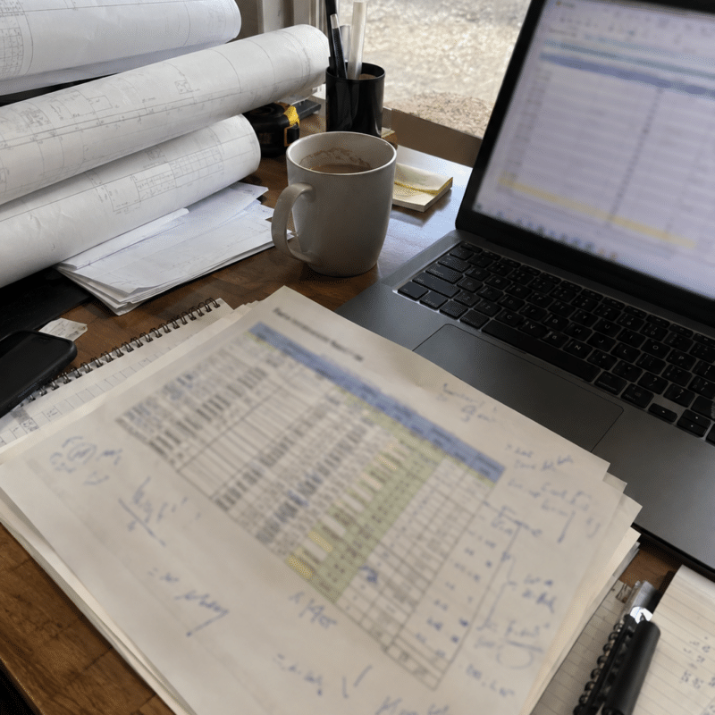Rolled construction drawings and a printed schedule on a desk beside a laptop showing a spreadsheet