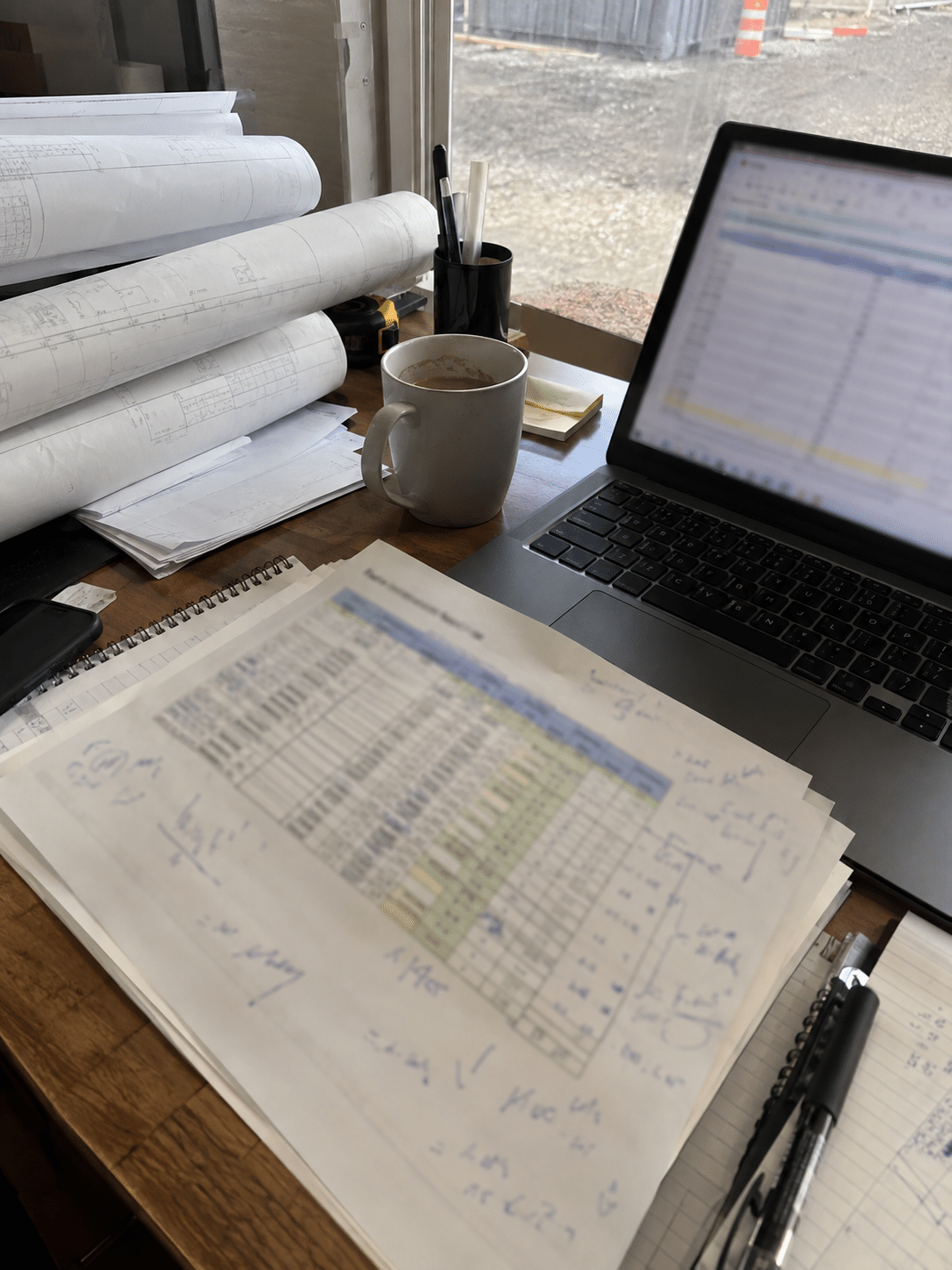 Rolled construction drawings and a printed schedule on a desk beside a laptop showing a spreadsheet